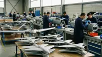 Atelier d'assemblage de drones militaires en France : fuselages gris alignés sur une chaîne, techniciens en blouse au travail, ailes volantes et munitions téléopérées au premier plan.