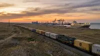 Train de conteneurs traversant la steppe kazakhe vers un port de la mer Caspienne, grues portuaires et navire roulier à l'horizon dans la lumière du soir.